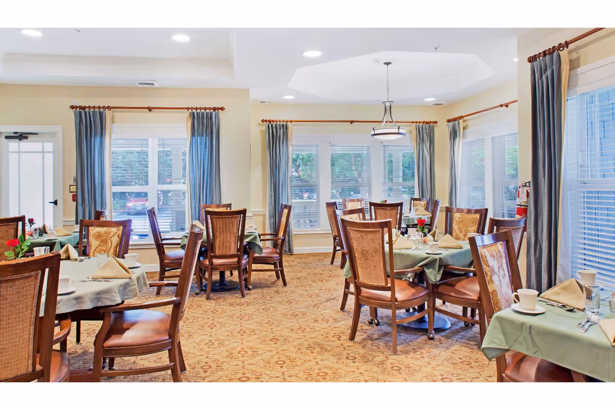 Bright dining room with multiple round tables set for meals, wooden chairs, large windows with curtains, and a patterned carpet.