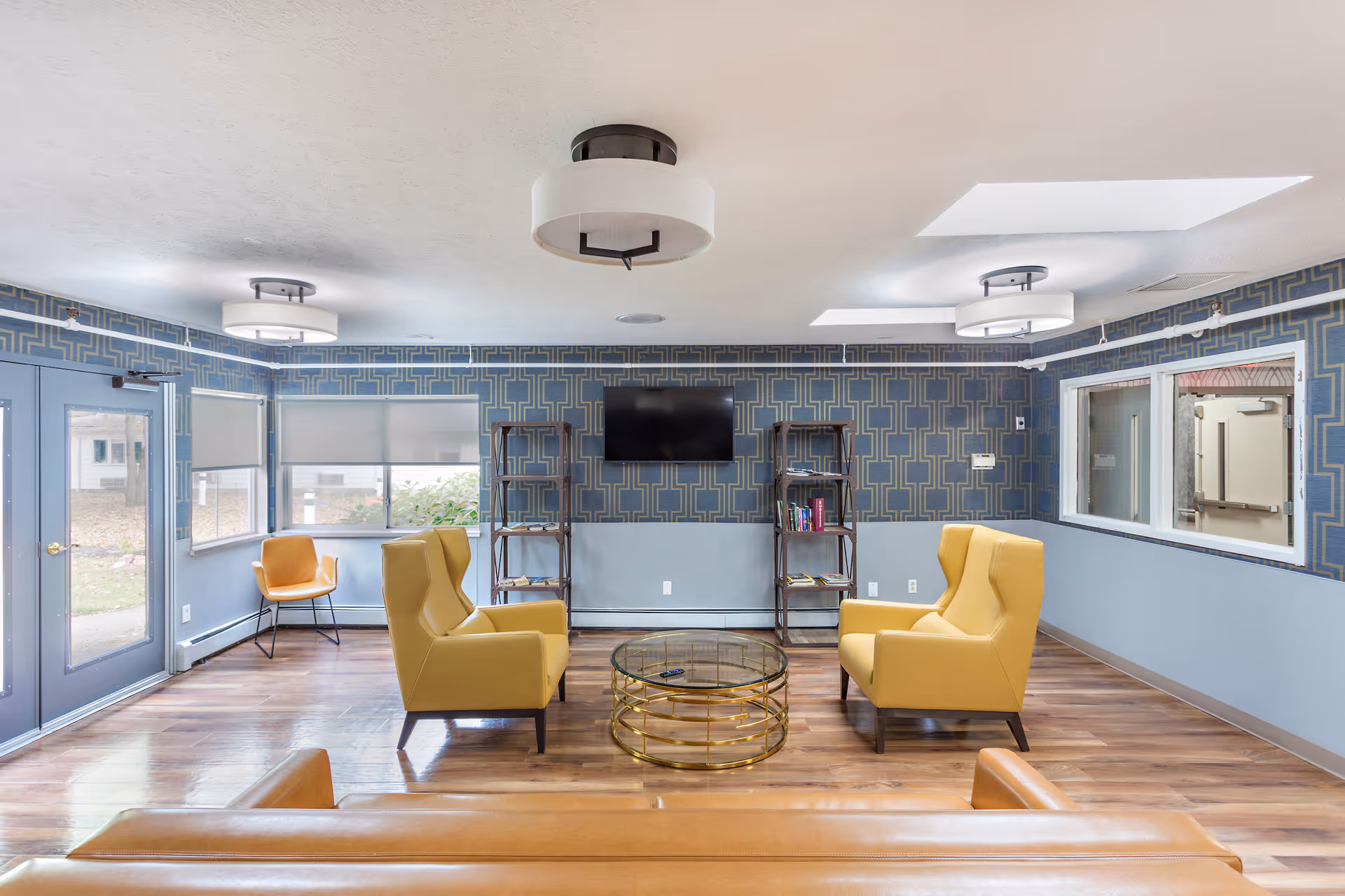 Bright seating lounge with yellow armchairs around a glass coffee table facing a wall-mounted TV and bookshelves.