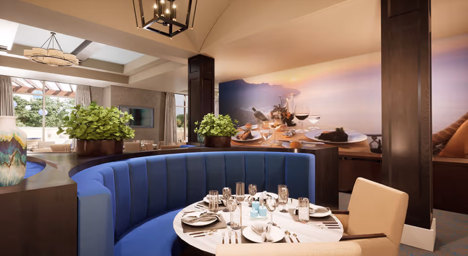 A dining area with a round table set for a meal, featuring plates, glasses, and silverware. The seating includes a curved blue upholstered bench and a beige chair. There are two large potted plants on a dark wooden divider behind the bench. In the background, there is a large wall mural depicting a scenic sunset view with a table set with wine glasses and food. The room has large windows with curtains, allowing natural light to enter.