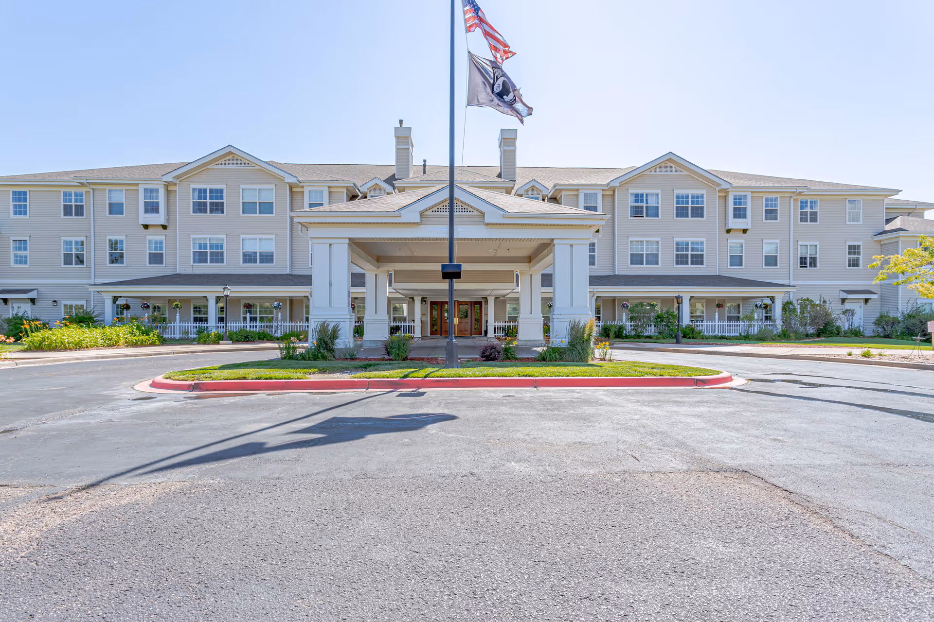 Front exterior view of Bear Creek Assisted & Senior Living facility showing a large three-story building with multiple windows, a covered entrance with white pillars, and two flags flying on a flagpole in front of the entrance.