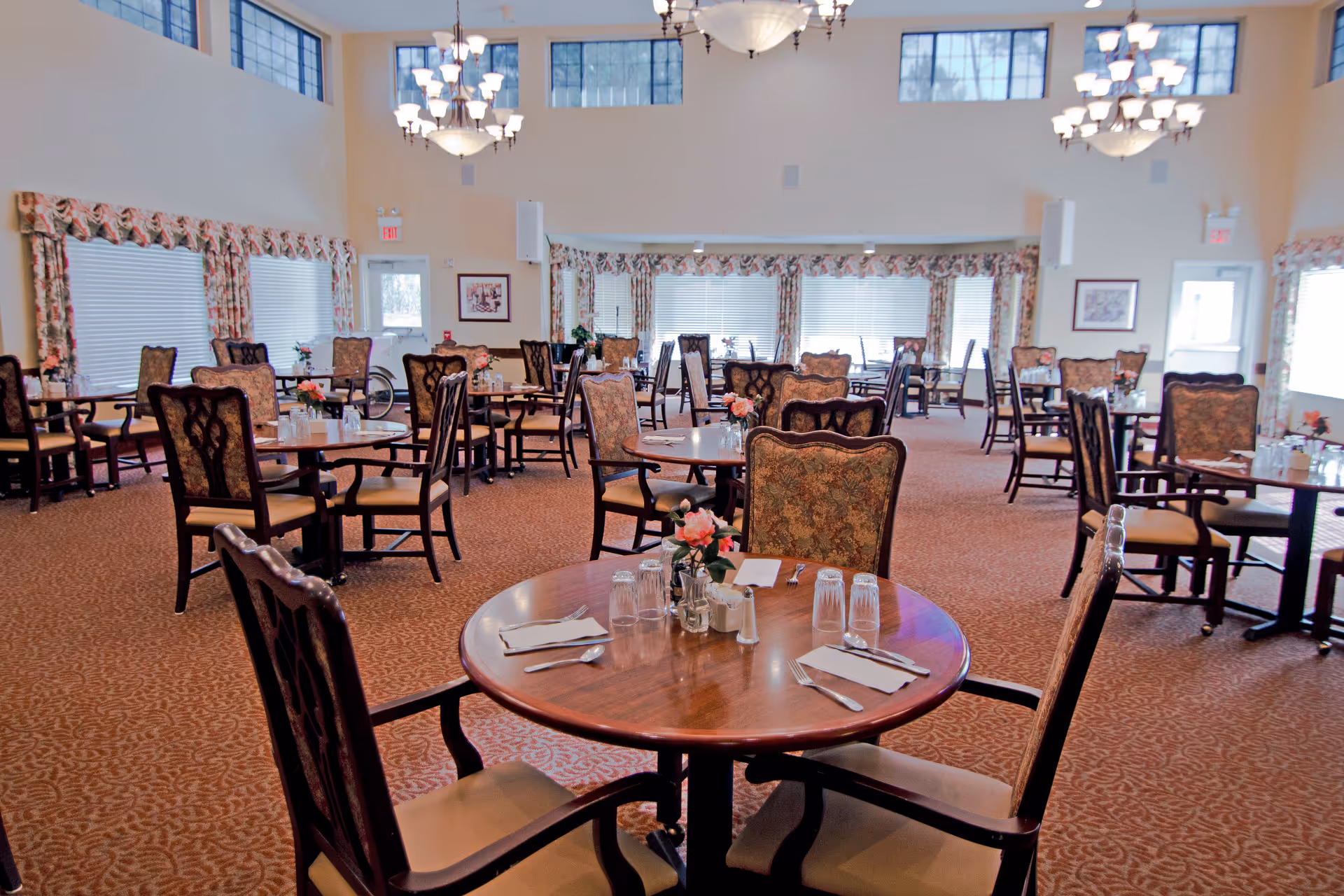 Spacious dining room with round tables and upholstered chairs set for meals under chandeliers.