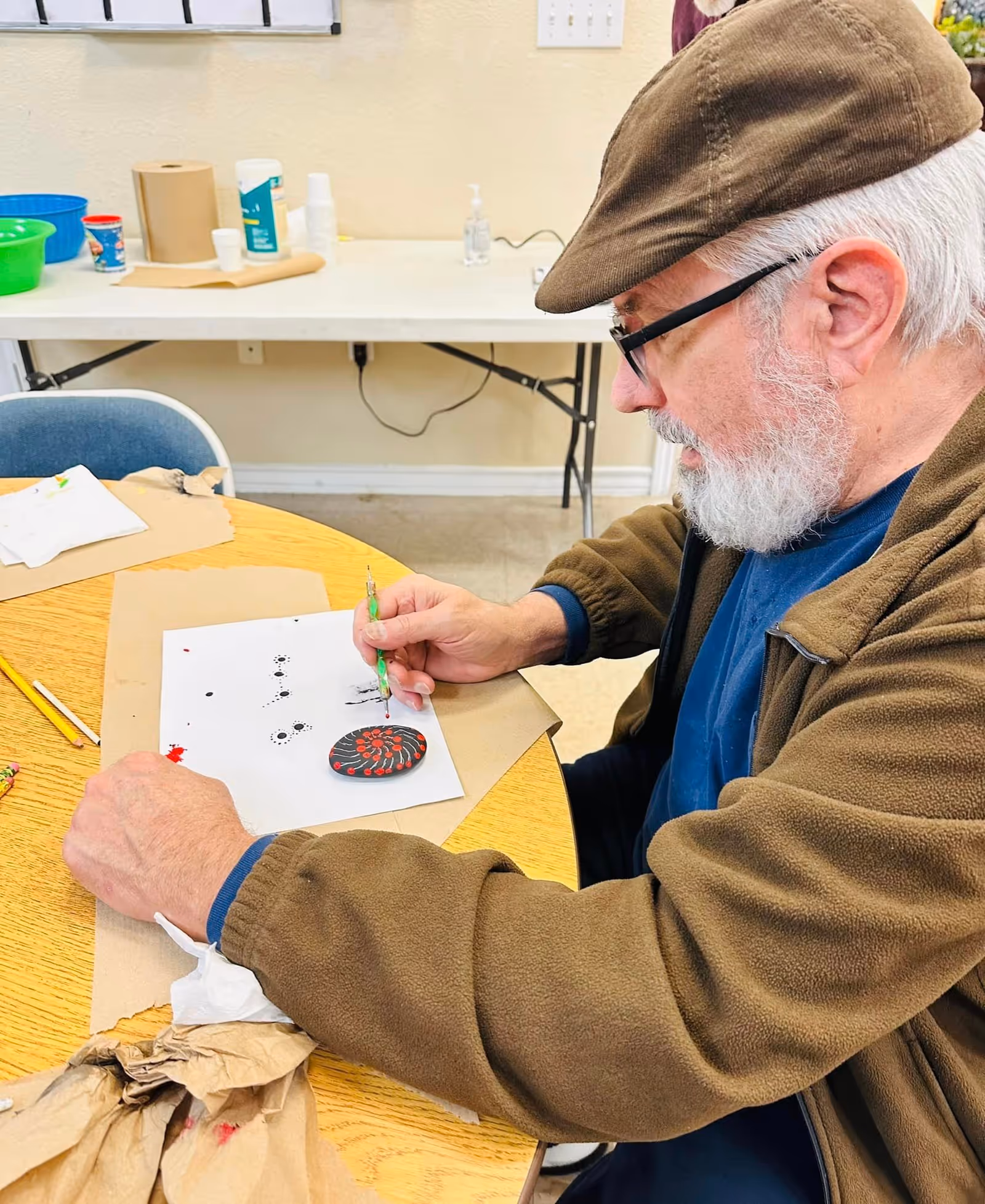 An elderly man with a white beard and glasses wearing a brown cap and jacket is sitting at a round wooden table, painting a small rock with red and black patterns. Various art supplies and paper towels are on the table, and a folding table with more supplies is in the background.
