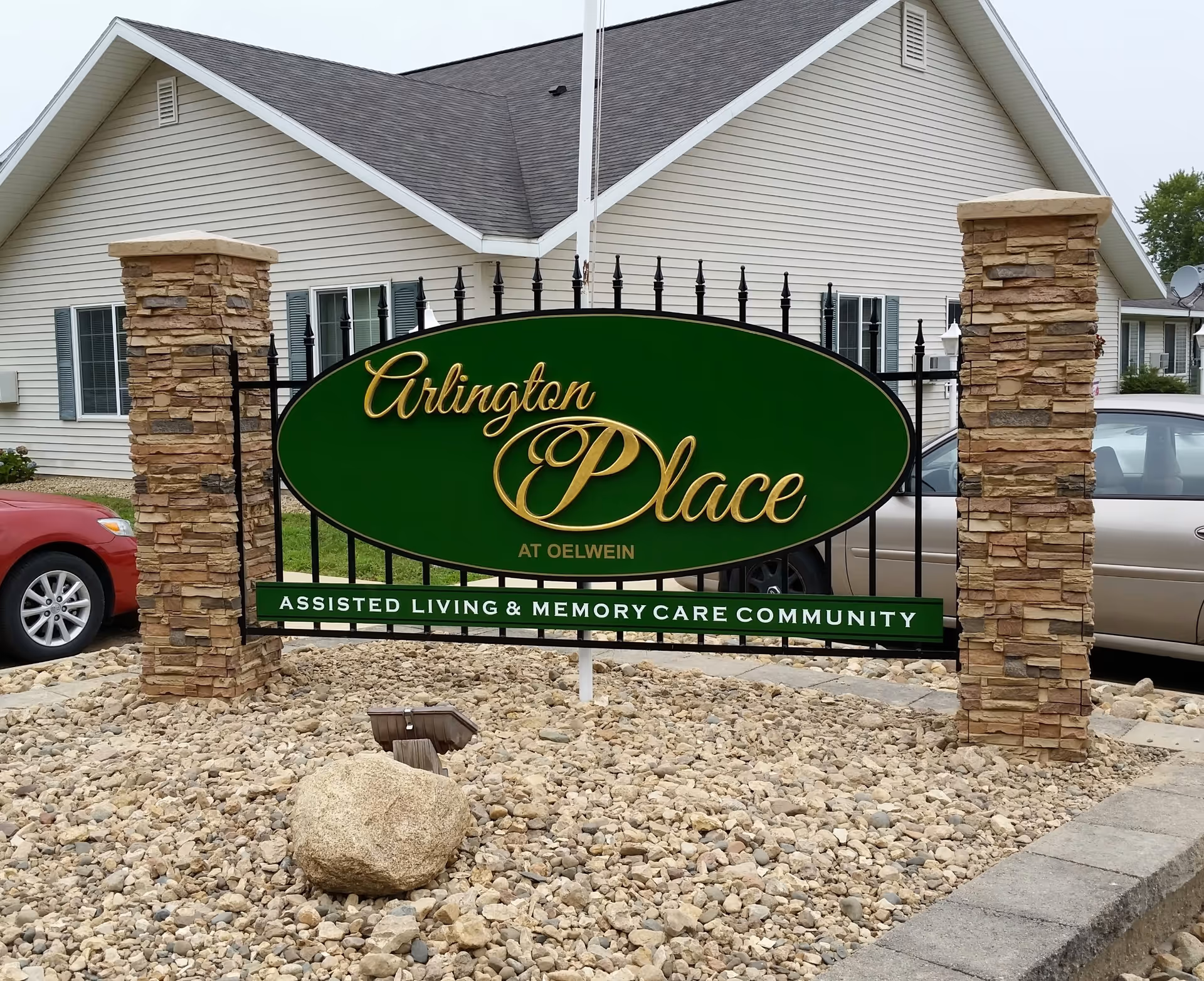 Green and gold sign reading 'Arlington Place at Oelwein Assisted Living & Memory Care Community' mounted on a black metal frame between two stone pillars, with a beige building and parked cars in the background.
