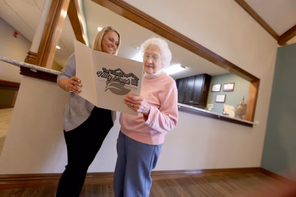 An elderly woman and a younger woman standing together inside a senior living facility. The elderly woman is holding and looking at a folder with the logo 'The Villas of Holly Brook' on it, while the younger woman smiles and looks on. They are in a hallway area with wooden flooring and a reception counter in the background.