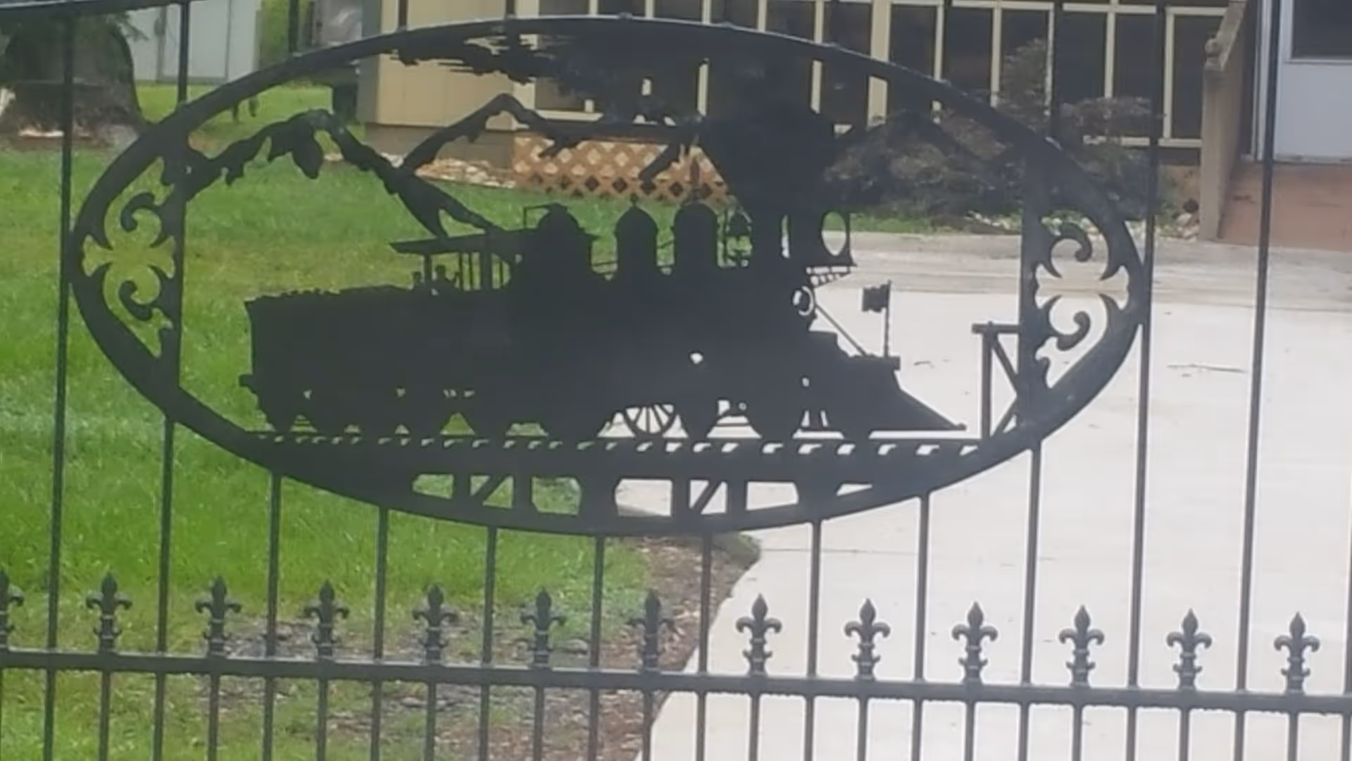 Black wrought iron gate with a decorative oval panel featuring a silhouette of a vintage steam locomotive train, set against a background of green grass and a concrete pathway leading to a building.