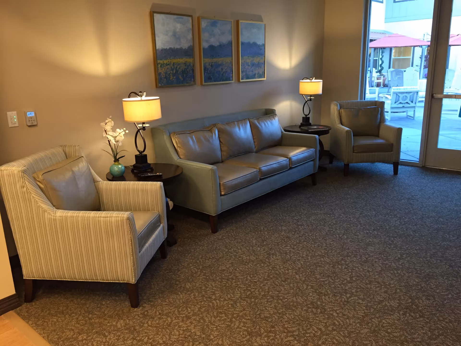 A cozy seating area with a leather sofa and two armchairs arranged around two small round tables with lamps. There are three framed landscape paintings on the wall above the sofa. A glass door leads to an outdoor patio with white chairs and red umbrellas.