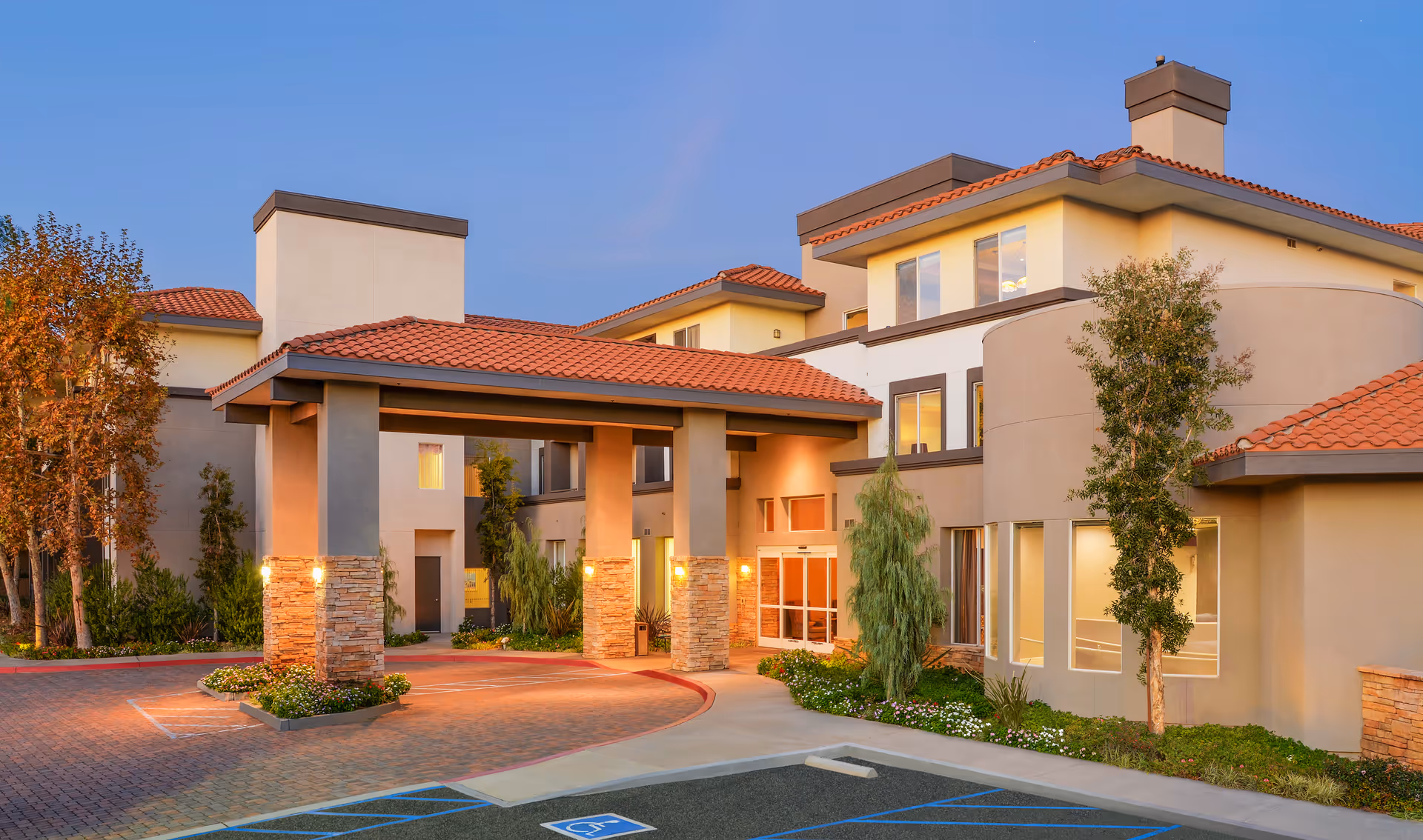 Exterior view of a senior living facility building with beige walls and red tiled roof, featuring a covered entrance supported by stone pillars, surrounded by landscaped greenery and a parking area with a handicapped parking space.