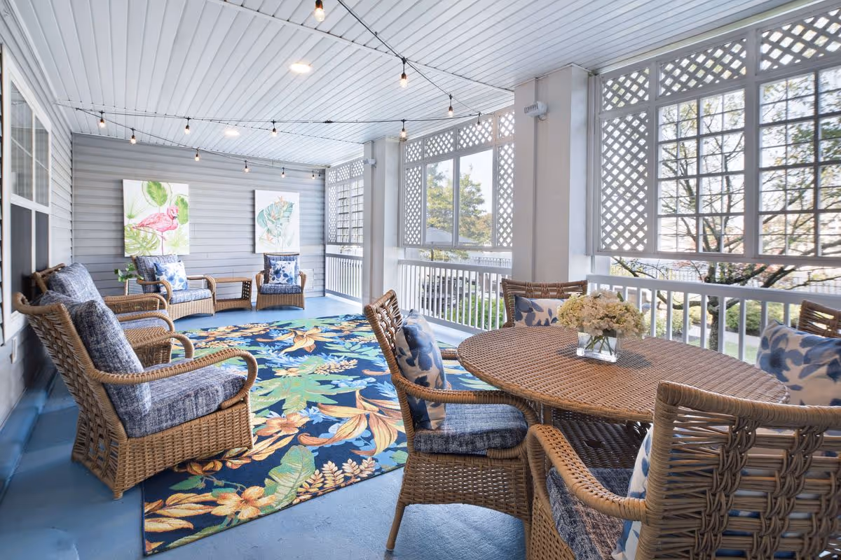 A bright and airy enclosed porch area with wicker furniture including a round table with four chairs and a seating area with two armchairs and a loveseat. The floor is covered with a colorful floral rug, and the walls have two framed tropical bird paintings. String lights hang from the ceiling, and large windows with lattice panels let in natural light and offer a view of trees outside.