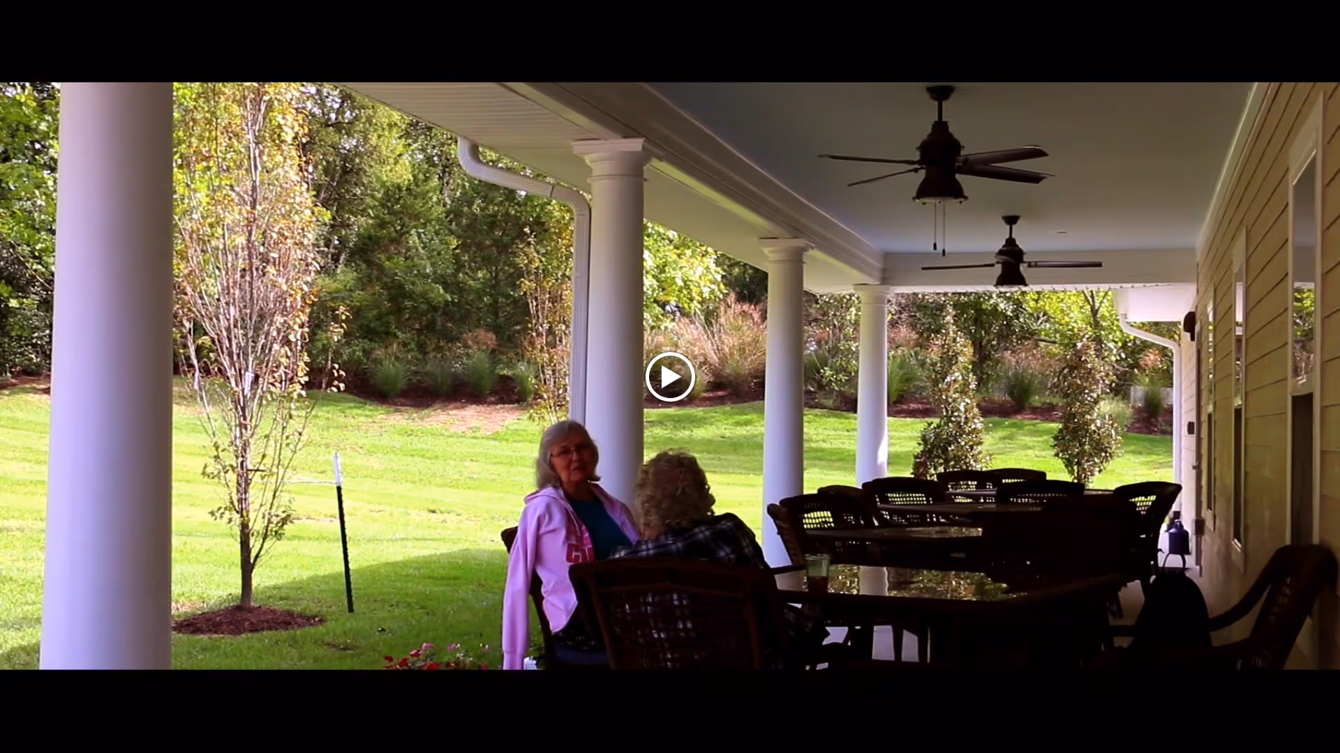 Two elderly women sitting and conversing on a covered outdoor patio with white columns, ceiling fans, and several tables and chairs. The patio overlooks a green lawn with trees and shrubs in the background.
