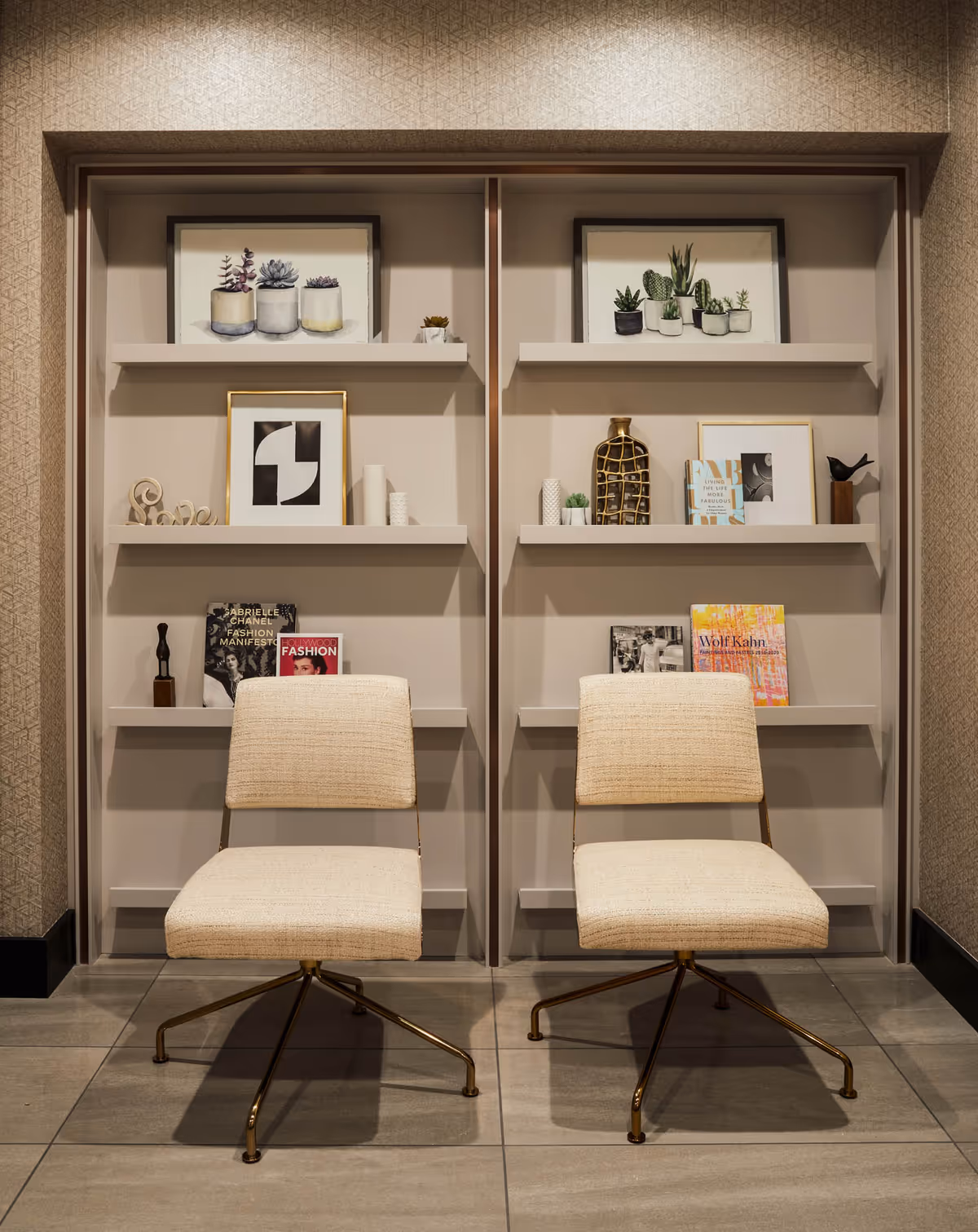 Two beige upholstered chairs with metal legs are placed in front of a built-in shelving unit. The shelves hold framed botanical prints, small decorative objects, books, and magazines. The setting appears to be a modern, stylish interior space with neutral tones and tiled flooring.