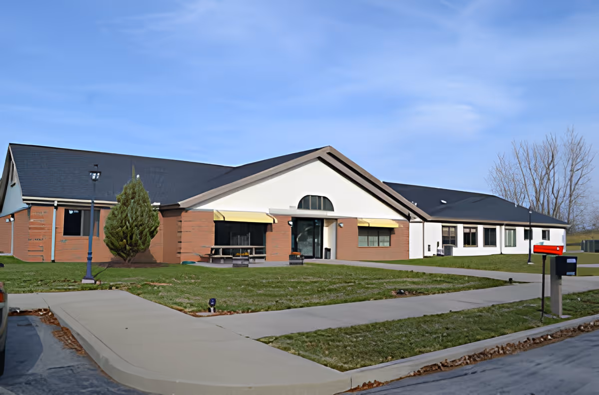 Exterior view of a single-story building with a black roof, white and brick walls, yellow awnings over windows, a sidewalk leading to the entrance, a red mailbox, and a small tree on the lawn under a clear blue sky.