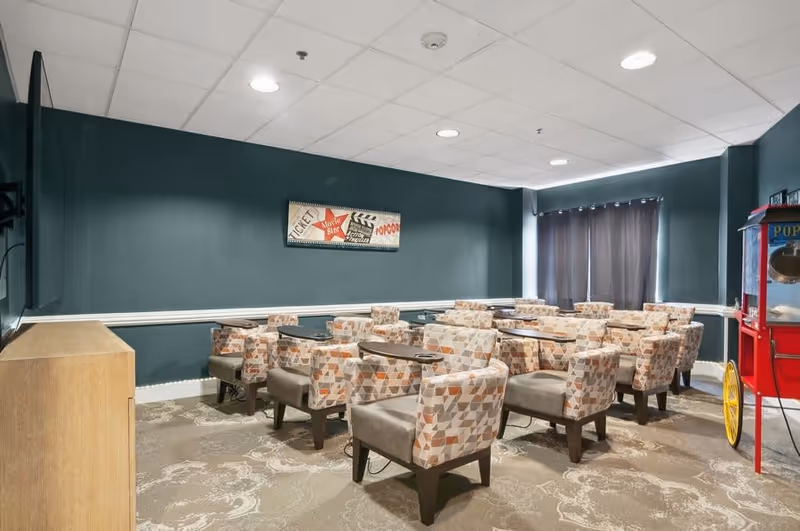 A small theater-style room with several upholstered chairs arranged in rows, each chair equipped with a small attached table. The walls are painted dark teal, and there is a movie-themed wall decoration. A red popcorn machine is visible on the right side of the room, and a large TV screen is mounted on the left wall.