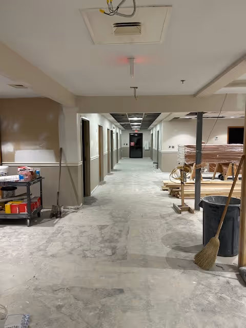 A long indoor hallway under construction or renovation with unfinished flooring and ceiling panels. Construction materials, tools, and equipment are scattered along the sides, including a broom, trash can, and a cart with supplies. The walls are painted beige and white, and several doorways line the hallway.