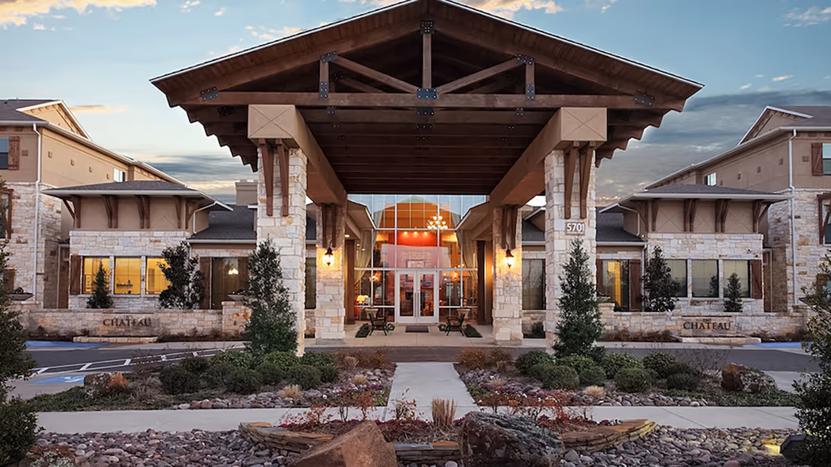 Front entrance of a stone-faced senior living building with a large wooden portico, glass doors, and landscaped driveway.