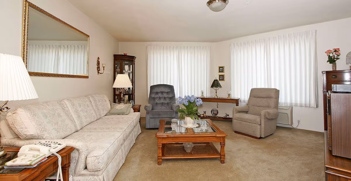Bright furnished living room with a long sofa, two recliners, a wooden coffee table and lamps in front of curtained windows.