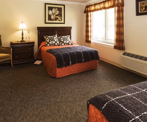 A cozy bedroom in an assisted living facility featuring two beds with orange bedspreads and dark patterned throws. There is a wooden nightstand with a lamp and decorative items between the beds. The room has a large window with plaid curtains allowing natural light to enter, and a framed floral artwork hangs above one bed. The floor is carpeted, and there is a heating/cooling unit beneath the window.