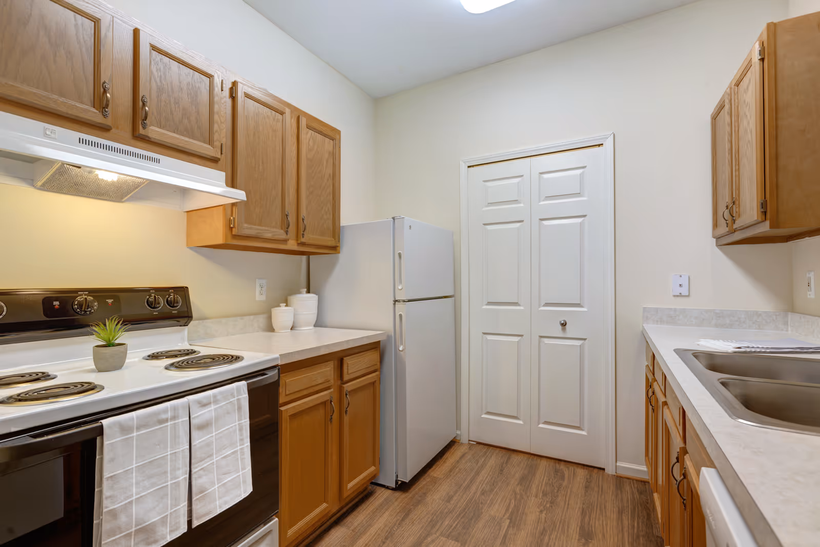 A compact kitchen with wooden cabinets, a white refrigerator, an electric stove with a white range hood, and a double sink. The countertops are light-colored, and the floor has a wood finish. There are two white towels hanging on the oven handle and a small potted plant on the stove.