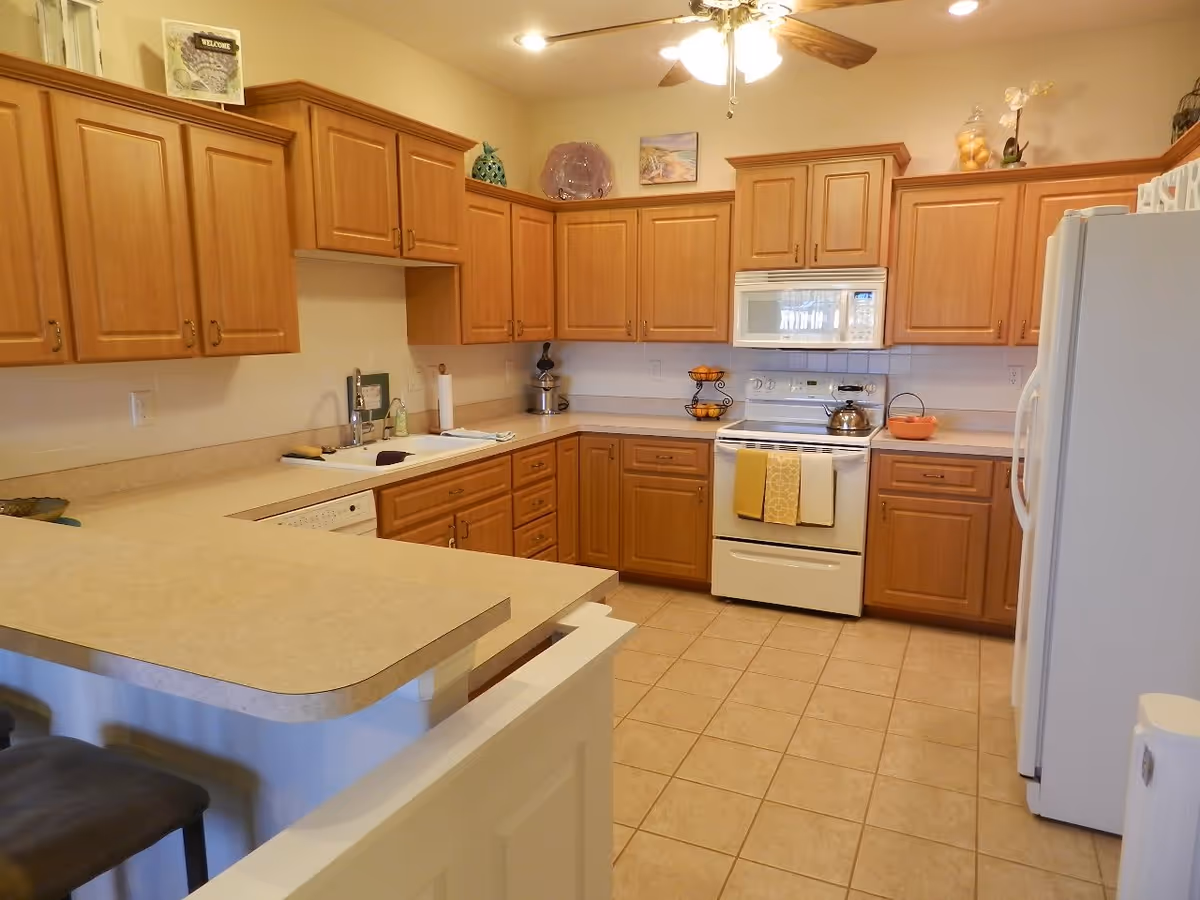 A spacious kitchen with wooden cabinets, beige countertops, and tiled floor. The kitchen features a white stove with a microwave above it, a white refrigerator, a sink, and a ceiling fan with wooden blades. There are two bar stools at the counter and various decorative items on top of the cabinets.