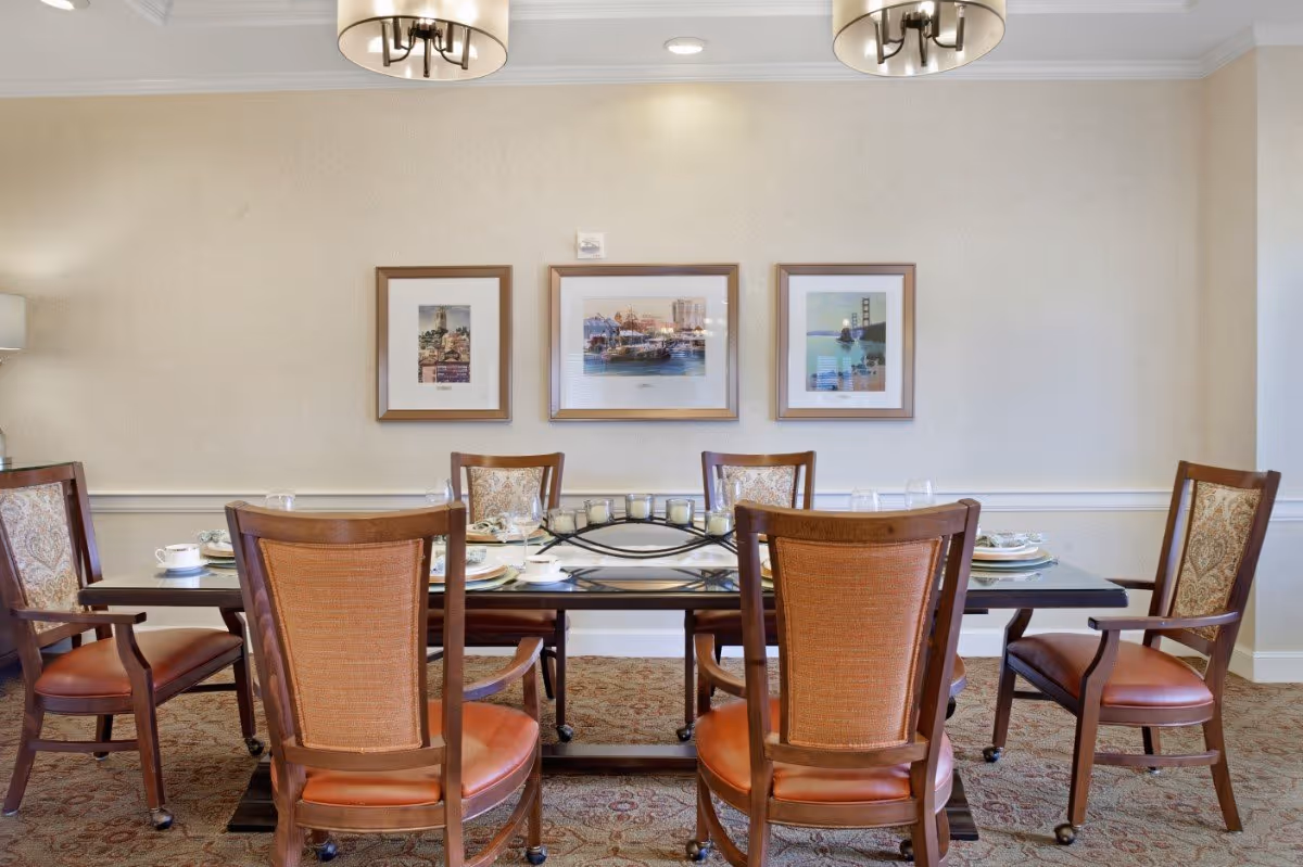 A dining room with a rectangular wooden table set for six people. The chairs have wooden frames with cushioned seats and backs. The table is set with plates, cups, and glasses. Three framed pictures hang on the beige wall behind the table. Two ceiling light fixtures are visible above the table.
