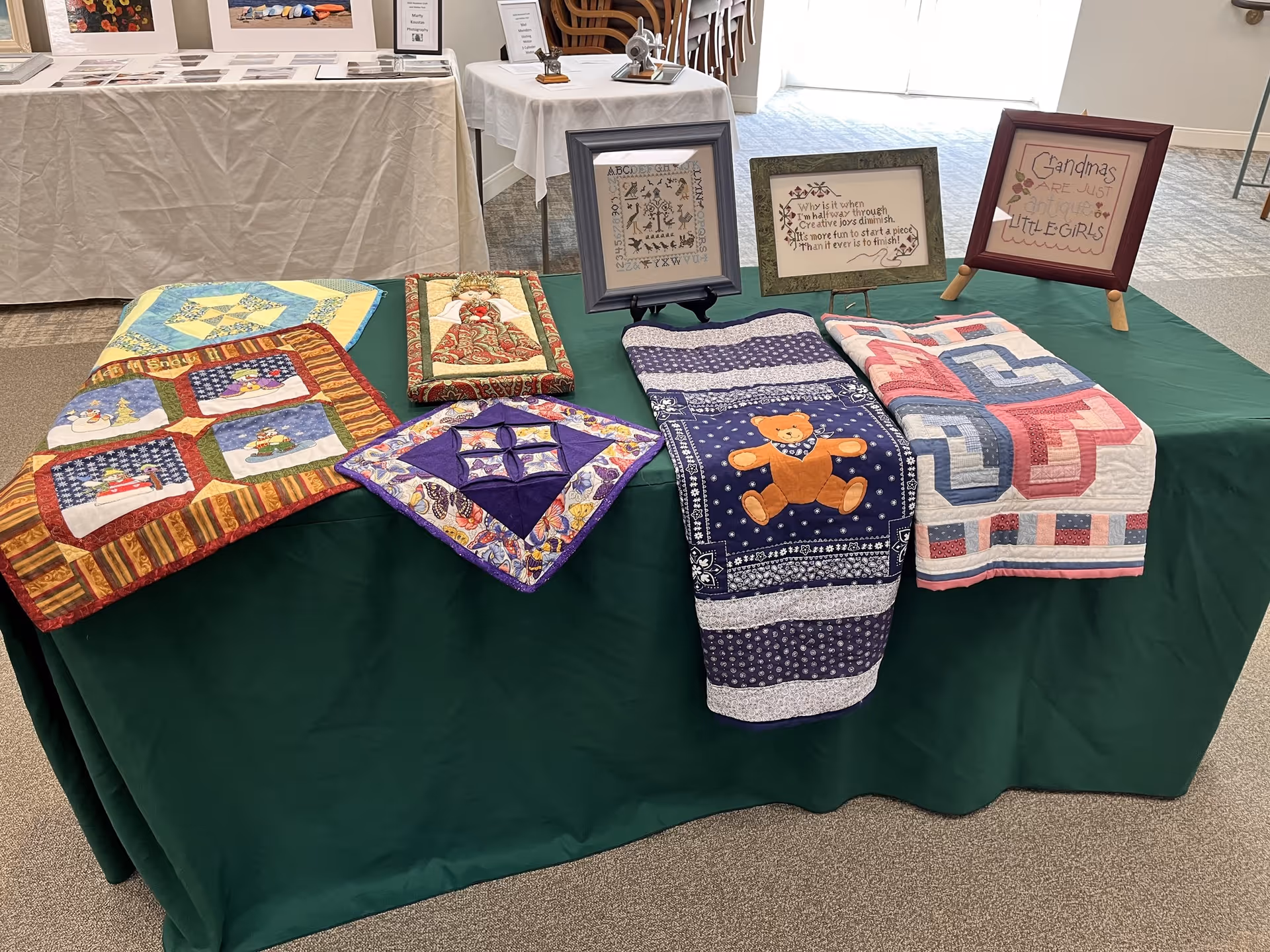 A display table covered with a green cloth showcasing various handmade quilts and framed cross-stitch art pieces. The quilts feature different patterns and colors, including a teddy bear design and geometric shapes. Behind the table, there is another table with a white cloth and additional framed artwork and items.