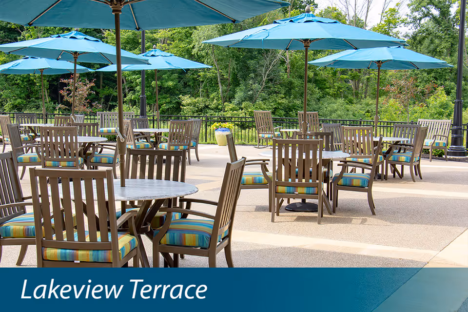Outdoor patio area with multiple round tables and wooden chairs with striped cushions, shaded by large blue umbrellas. Green trees and foliage are visible in the background.