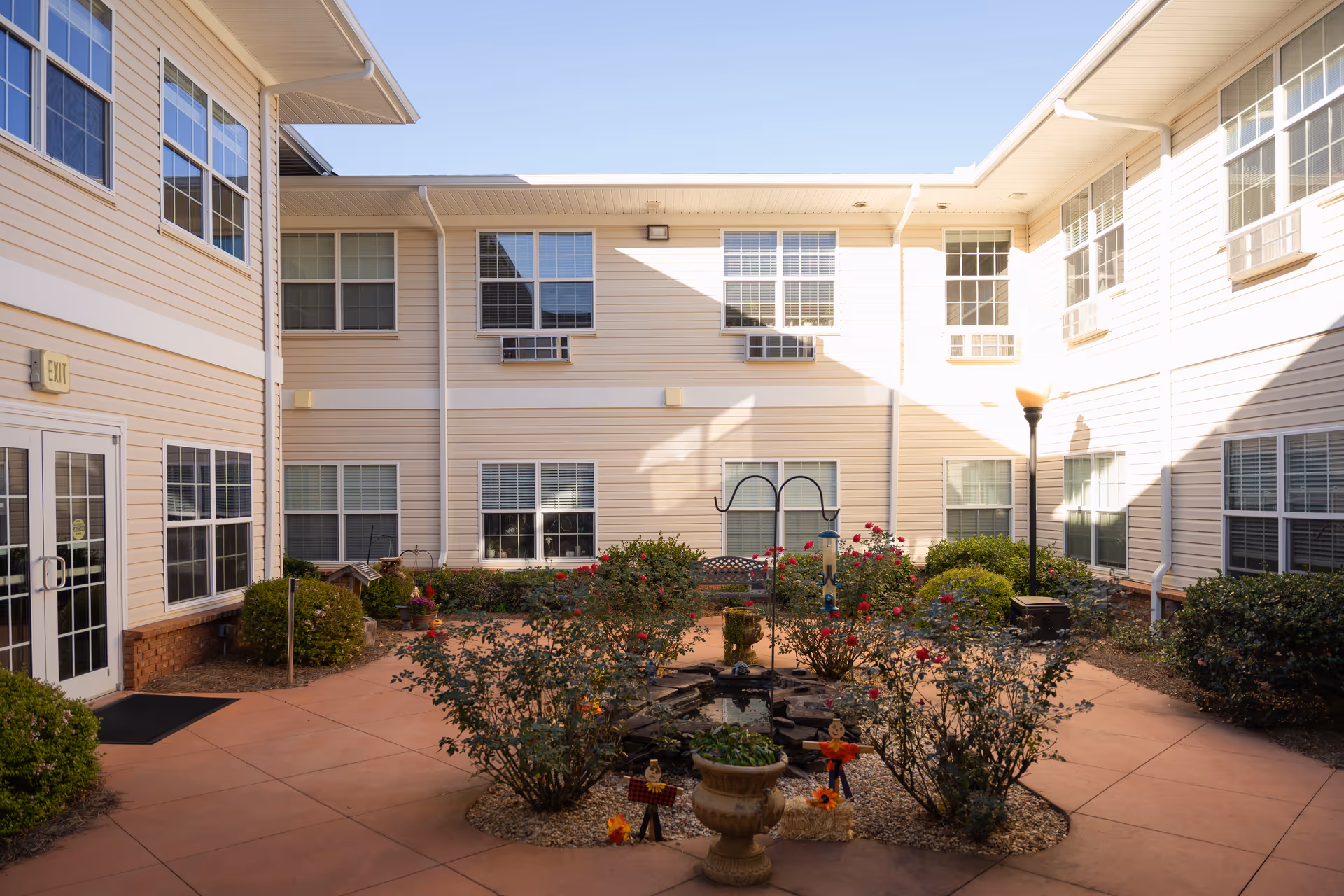 Sunny landscaped interior courtyard with planted beds, a small fountain, benches and the surrounding two-story beige senior living building.