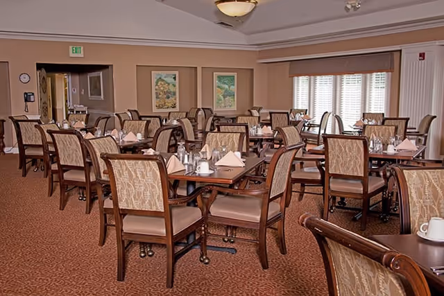 Dining room with multiple wooden tables and upholstered chairs set with napkins and place settings.