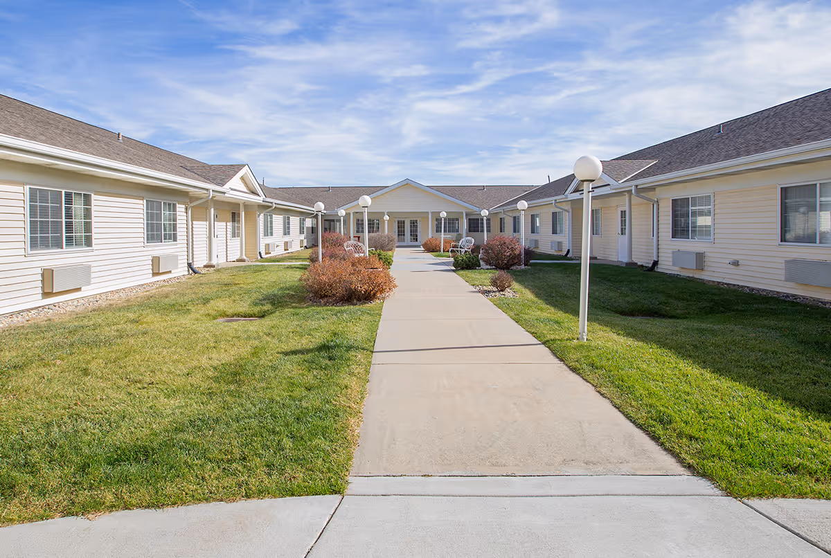 A wide concrete pathway leads through a grassy courtyard area with bushes and white globe lamp posts on either side, flanked by single-story beige buildings with multiple windows under a partly cloudy blue sky.