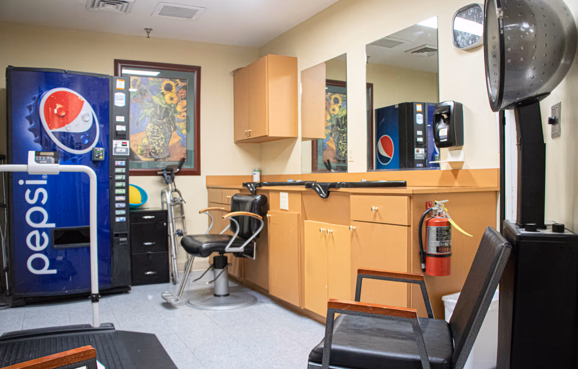 Interior room with a Pepsi vending machine, a black salon chair, a black waiting chair, a wall-mounted hair dryer, a fire extinguisher, wooden cabinets with mirrors, and a framed picture of sunflowers on the wall.