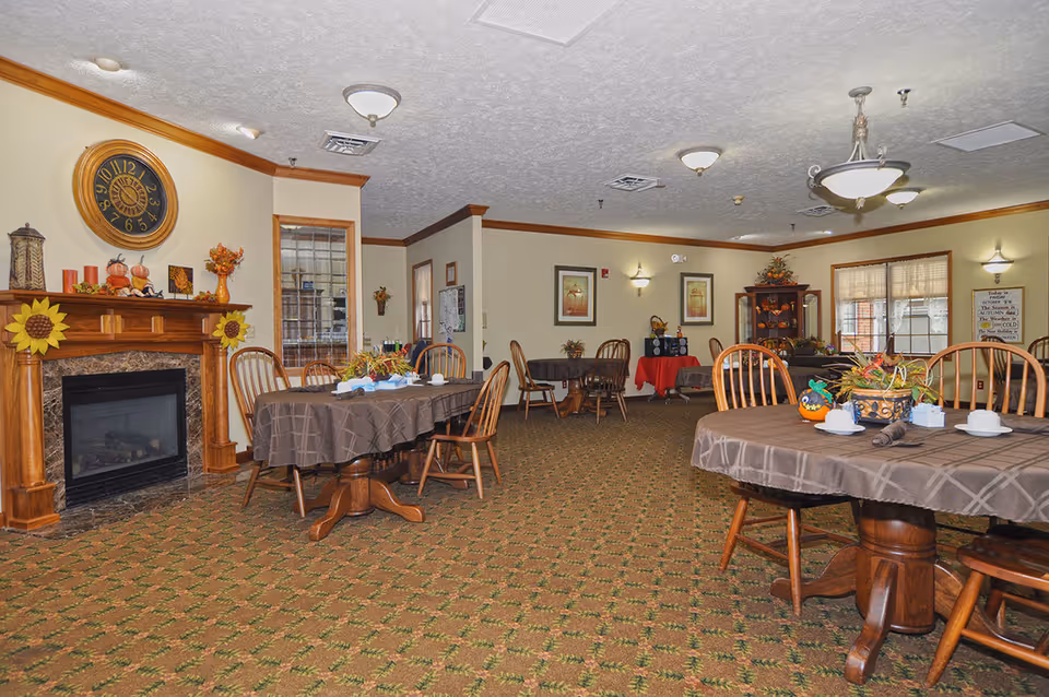 A cozy dining room in Magnolia Terrace Assisted Living featuring round tables covered with brown tablecloths, wooden chairs, a fireplace decorated with autumn-themed items, a large wall clock, framed artwork, and a carpeted floor with a patterned design.