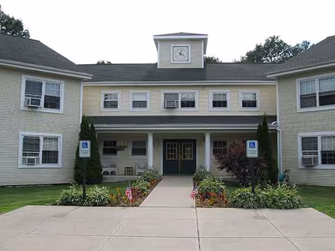 Front exterior of a yellowish senior housing building with a central entrance and landscaped walkway.