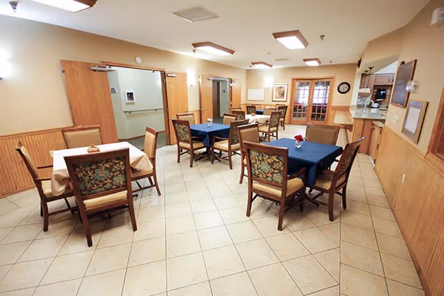 A dining room in a senior living facility with several tables covered with tablecloths, some beige and some dark blue. Each table is surrounded by wooden chairs with floral upholstery. The room has tiled floors, wood-paneled lower walls, and beige upper walls. There are ceiling lights and a serving window on the right side of the room.