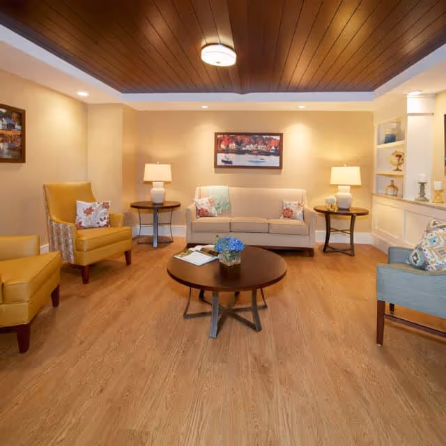 A cozy senior living room with a beige sofa, two yellow armchairs, and a light blue armchair arranged around a round wooden coffee table with a small plant and magazines. Two side tables with lamps flank the sofa, and a framed painting hangs on the wall above it. The room features warm lighting, wooden flooring, and a wooden ceiling with a central light fixture.