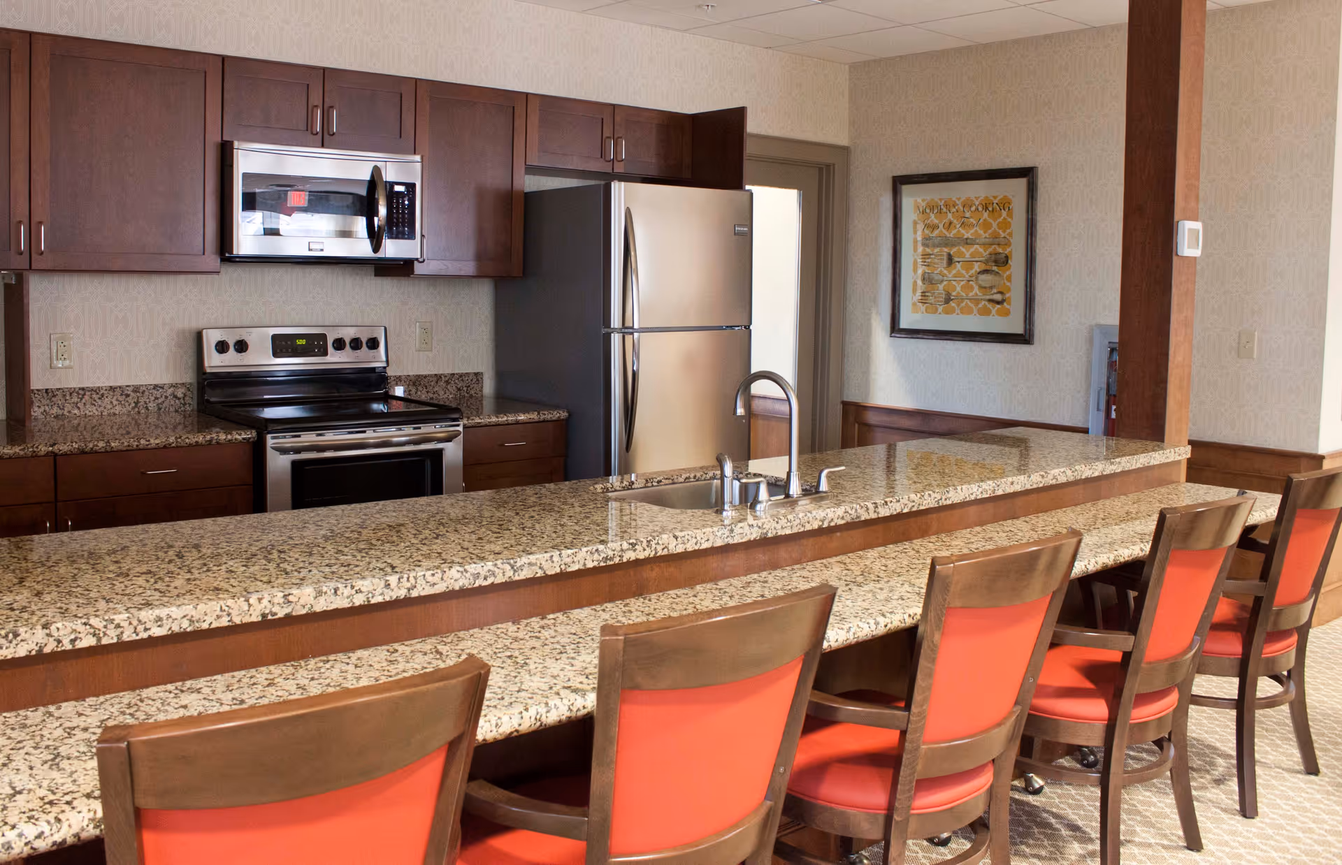 A modern kitchen area with a long granite countertop and a row of wooden chairs with red cushions. The kitchen features stainless steel appliances including a refrigerator, microwave, and stove. There is a sink with a faucet on the countertop and a framed artwork on the wall in the background.