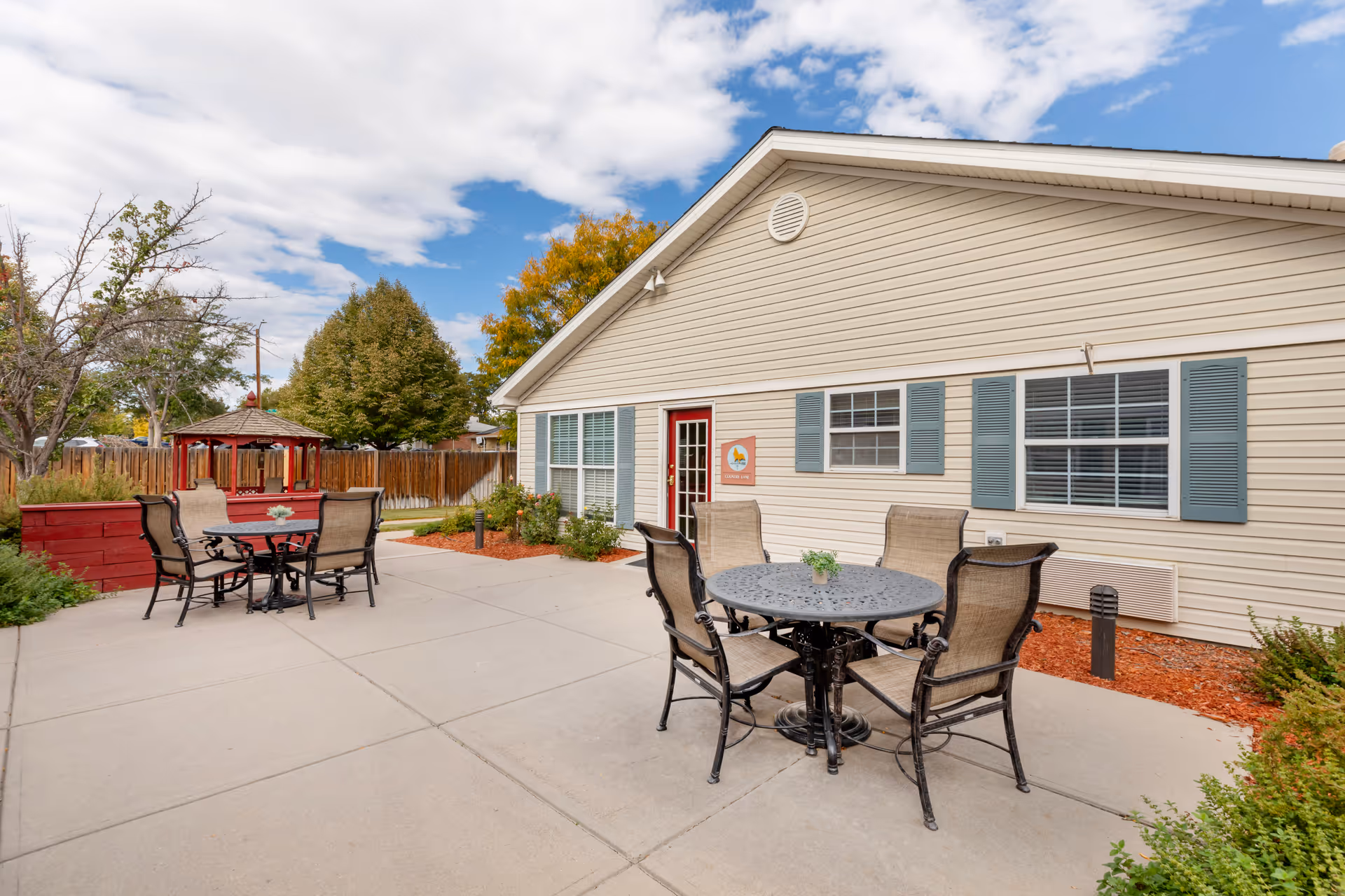Outdoor patio area at Brookdale Pinehurst Park with two round metal tables and several chairs on a concrete surface. The building has beige siding with blue shutters and a red door. There is a wooden gazebo and some trees and bushes in the background under a partly cloudy sky.