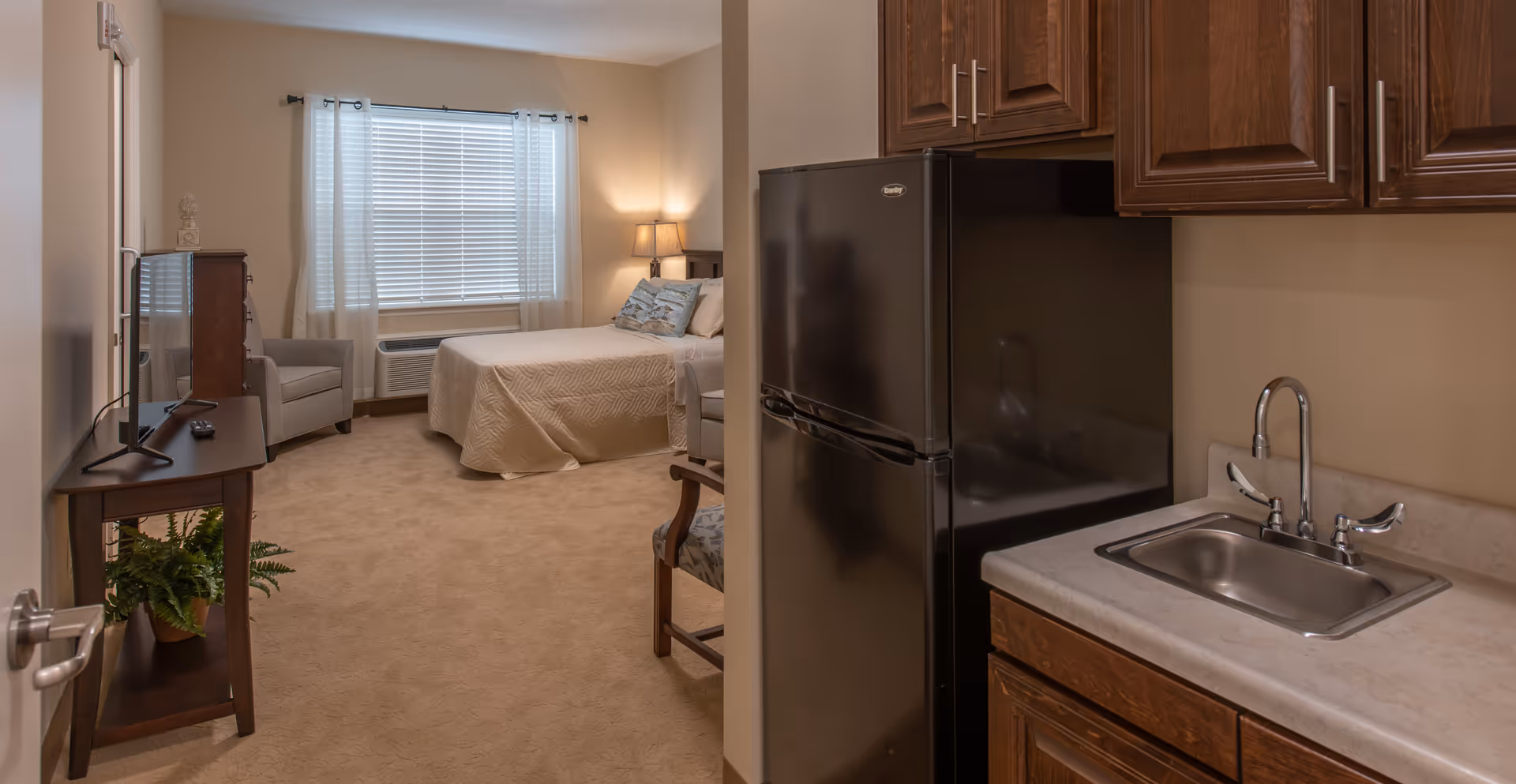 Interior view of a senior living facility room at The Addison of St. Johns showing a small kitchenette with a sink and refrigerator on the right, and a bedroom area with a bed, armchair, TV on a stand, and a window with blinds and curtains in the background.