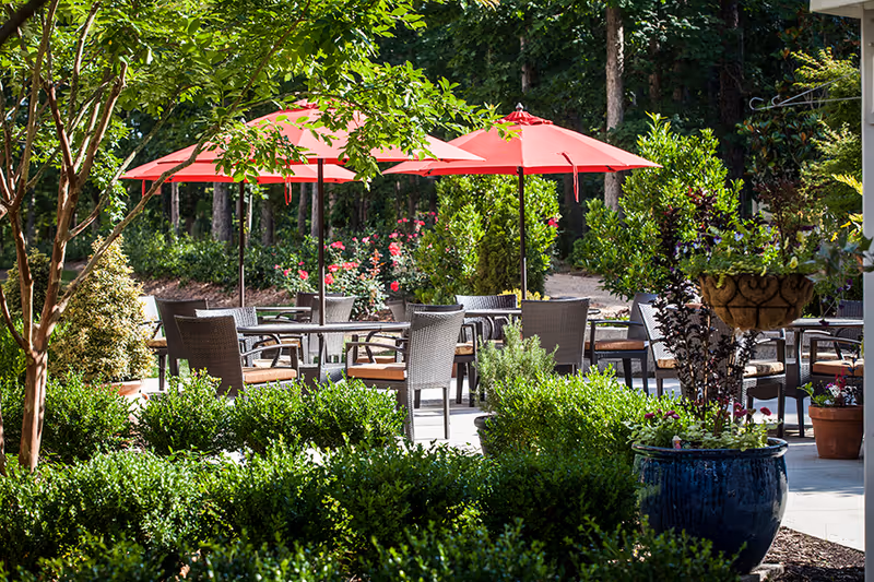 Outdoor patio with tables, wicker chairs and red umbrellas surrounded by greenery and potted plants.
