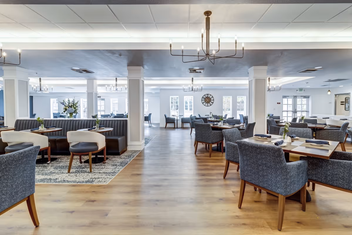 Spacious and well-lit dining room with multiple tables and chairs arranged neatly. The room features wooden flooring, modern chandeliers, large windows allowing natural light, and decorative plants on tables. There are cushioned booth seats along one side and comfortable armchairs around the tables.
