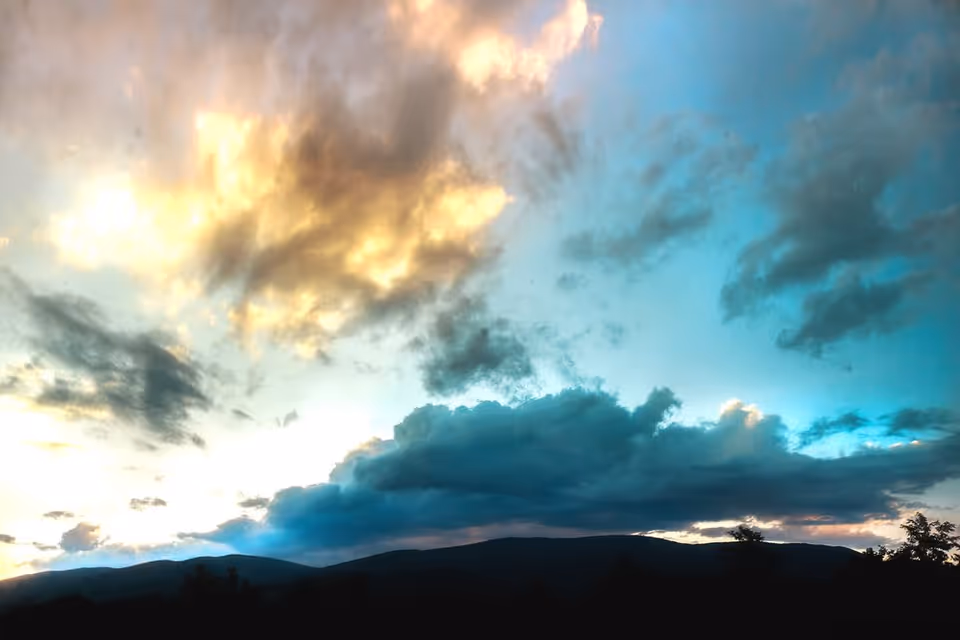 A scenic view of a mountain range silhouetted against a dramatic sky with clouds illuminated by the setting or rising sun, displaying shades of blue, orange, and yellow.