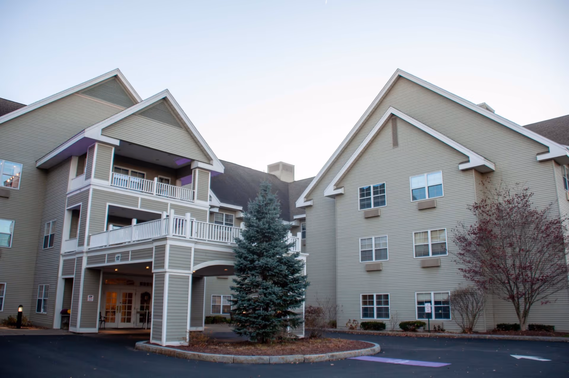 Three-story beige senior living building with a covered entrance, balconies, and a pine tree in front.