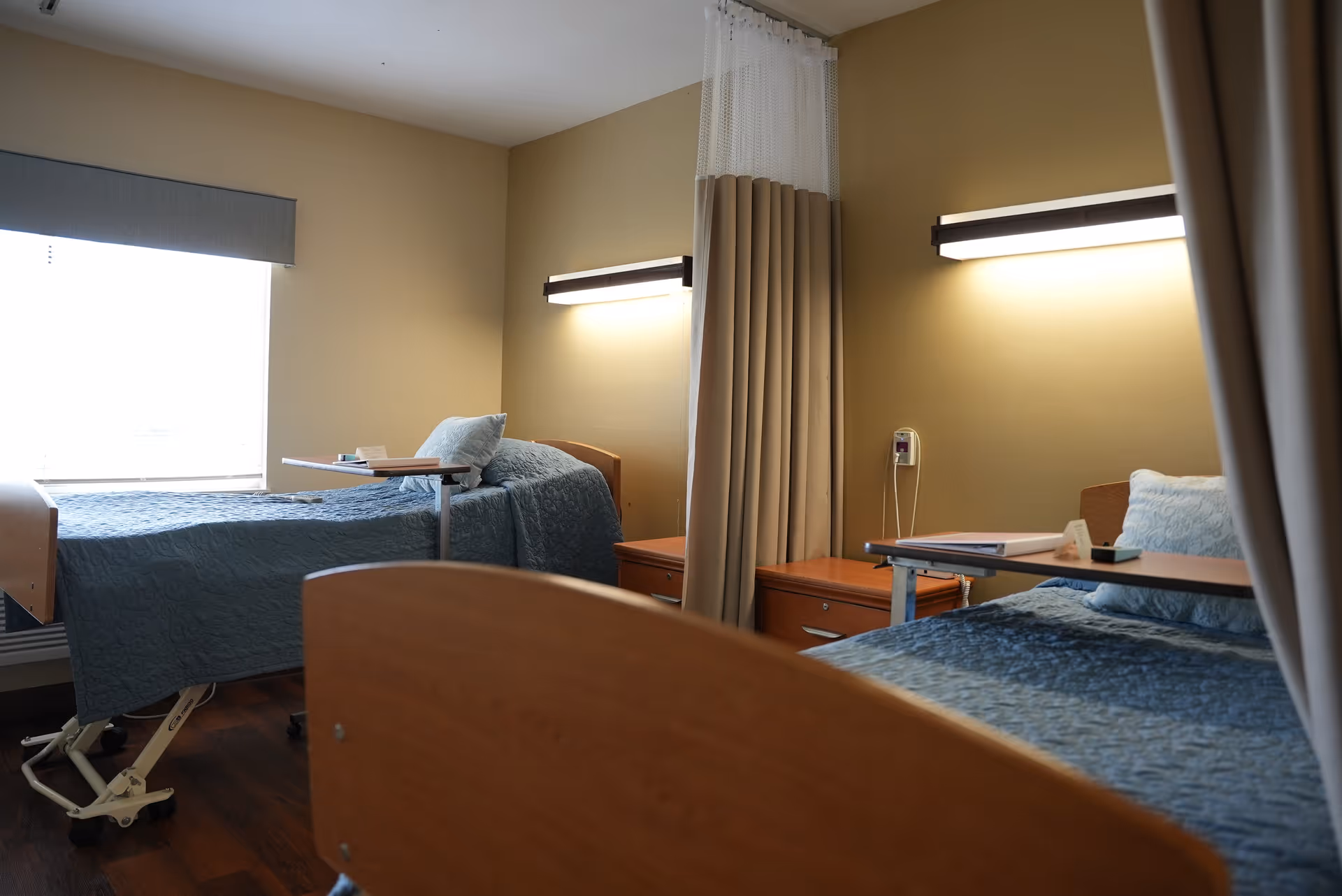 Two hospital-style beds with blue bedspreads separated by a privacy curtain in a senior care room with wall lights and bedside tables.