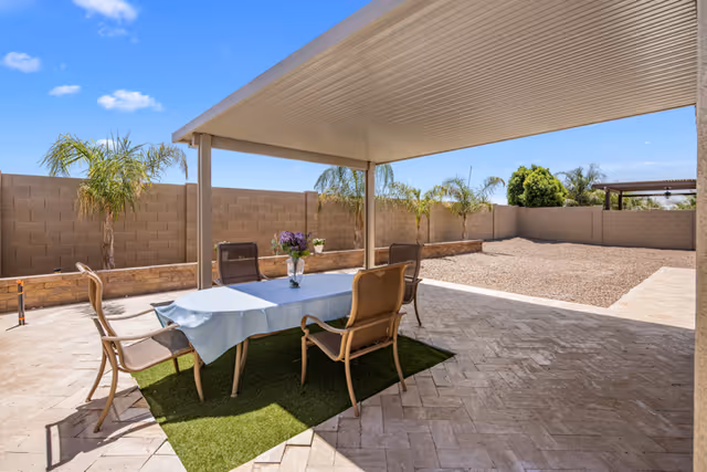 Outdoor patio area with a covered pergola, a table covered with a light blue tablecloth, four chairs around the table, and a vase with flowers on top. The patio is paved with stone tiles and has a small patch of artificial grass under the table. There are palm trees and a brick wall surrounding the yard under a clear blue sky.