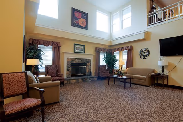 A spacious senior living facility common area with high ceilings and large windows allowing natural light. The room features a stone fireplace centered on one wall, flanked by two windows with maroon valances. There are multiple seating options including a beige sofa, two patterned armchairs, and a wooden chair with red upholstery. A coffee table with a floral arrangement sits in front of the sofa. A flat-screen TV is mounted on the wall to the right, and there are two table lamps on side tables. The carpet has a patterned design, and the walls are painted a warm beige color.