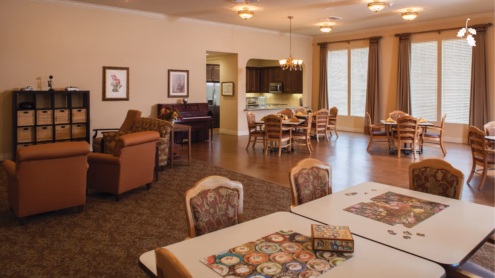 A spacious common area in a memory care community featuring a seating area with armchairs and a piano, several dining tables with chairs, large windows with blinds and curtains, and a kitchen area with dark wood cabinets and stainless steel appliances. Puzzle pieces and a puzzle box are on the tables in the foreground.