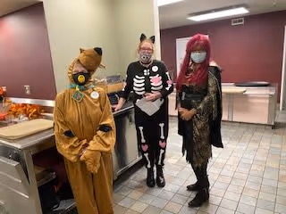 Three people dressed in Halloween costumes standing in a kitchen area. One person is wearing a Scooby-Doo costume, another is dressed in a black skeleton outfit with cat ears, and the third person is wearing a black and gold outfit with a red wig and face mask. The background shows kitchen counters and tiled flooring.
