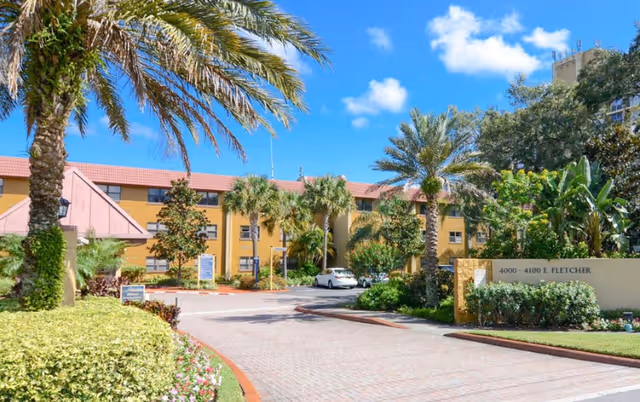 Exterior view of Concordia Village of Tampa showing a driveway lined with palm trees and landscaped greenery leading to a yellow multi-story building under a blue sky with scattered clouds.