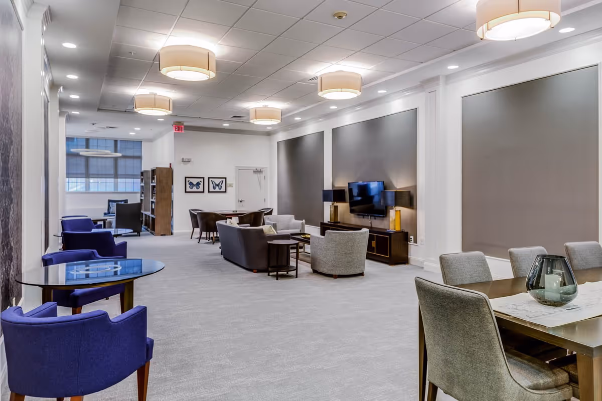 A spacious, modern common area with multiple seating arrangements including blue armchairs around a glass table, gray armchairs and sofas around a coffee table, and a dining table with gray chairs. The room features large gray wall panels, a mounted flat-screen TV, decorative lamps, and ceiling lights. Large windows allow natural light into the space.