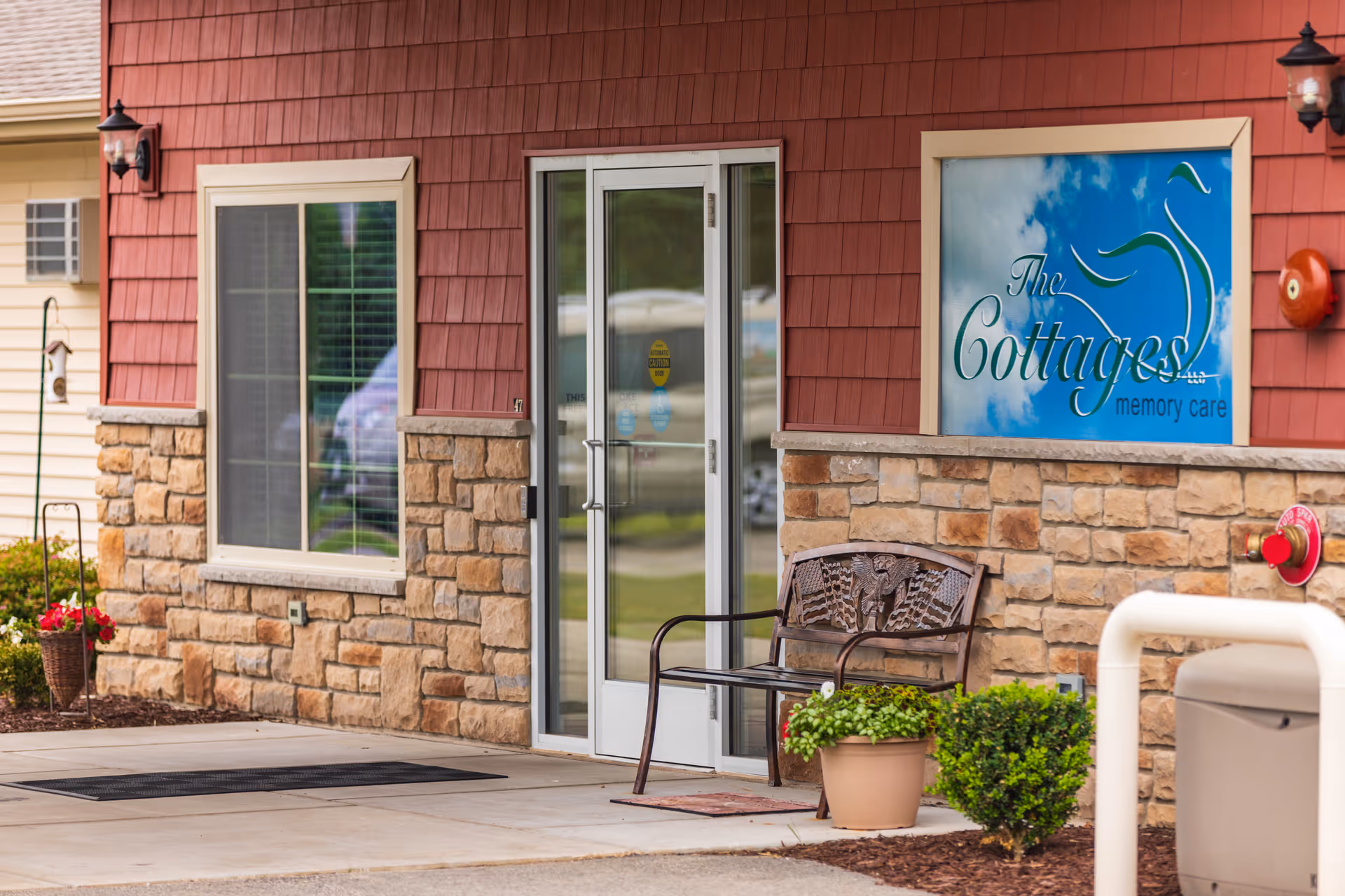 Front entrance of The Cottages memory care building with a glass door, stone facade, bench, potted plants, and a blue sign reading 'The Cottages'.