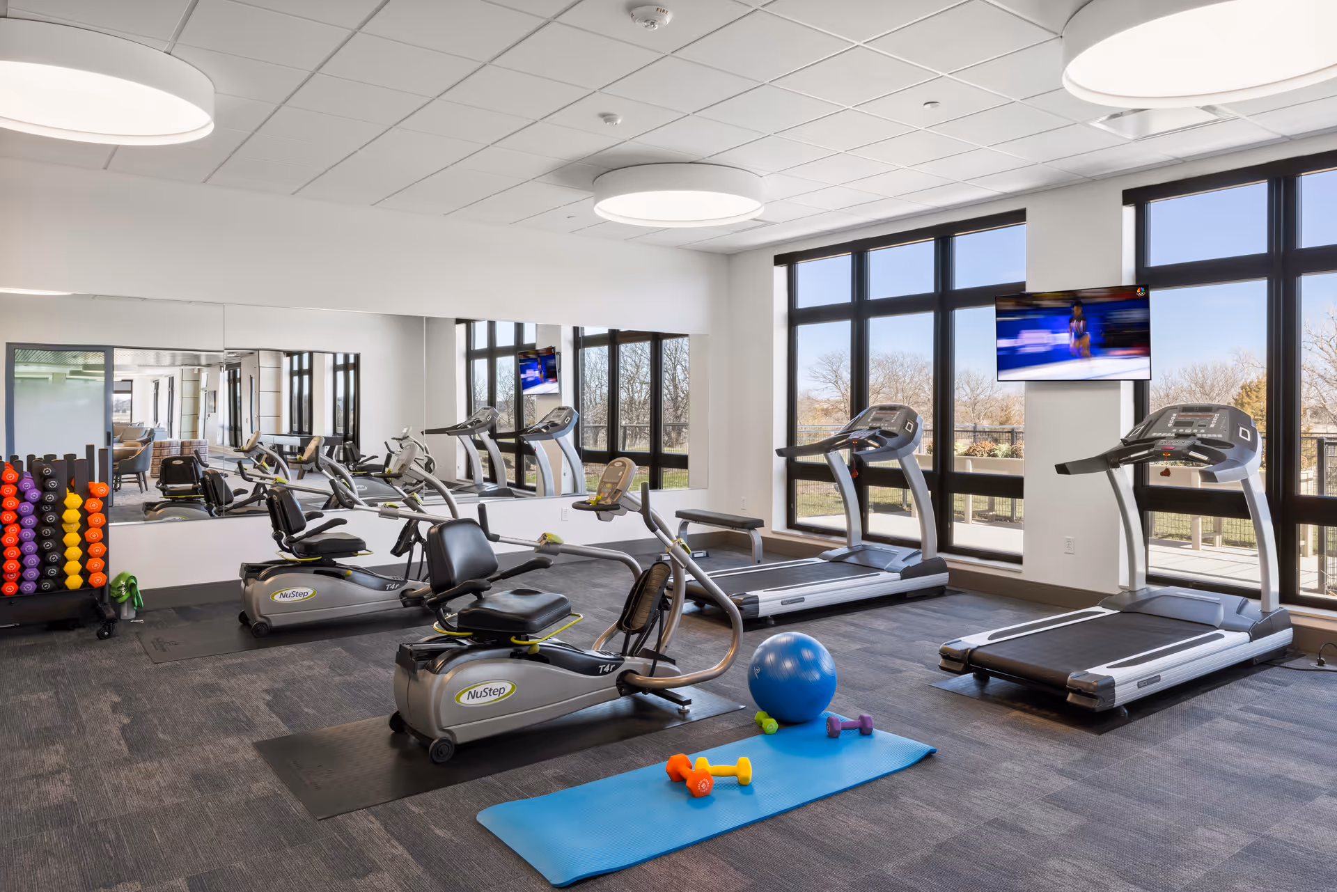 A bright and modern fitness room with large windows letting in natural light. The room contains two treadmills, two NuStep recumbent cross trainers, a rack of colorful dumbbells, a blue exercise ball, and a blue yoga mat with small dumbbells on it. A large mirror covers one wall, reflecting the equipment and windows. A flat-screen TV is mounted on the wall between the windows.