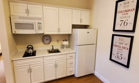Small kitchen area with white cabinets, a white refrigerator, a microwave, a kettle on the stovetop, a small sink, and two framed coffee-themed prints on the wall.